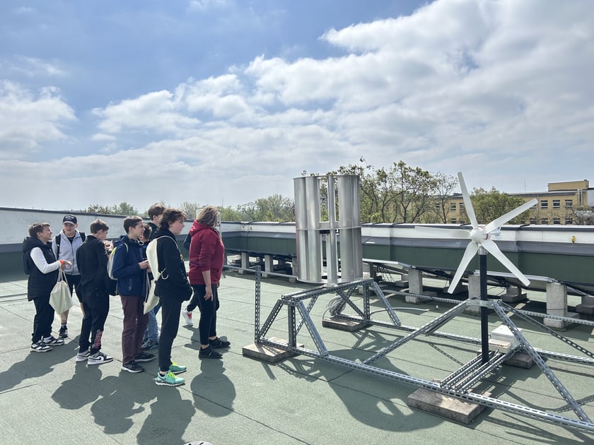 Grupa nastolatków stoi na dachu budynku i ogląda instalacje odnawialnych źródeł energii – pionową turbinę wiatrową oraz turbinę wiatrową z białymi łopatami, ustawione na metalowych konstrukcjach obok paneli fotowoltaicznych.