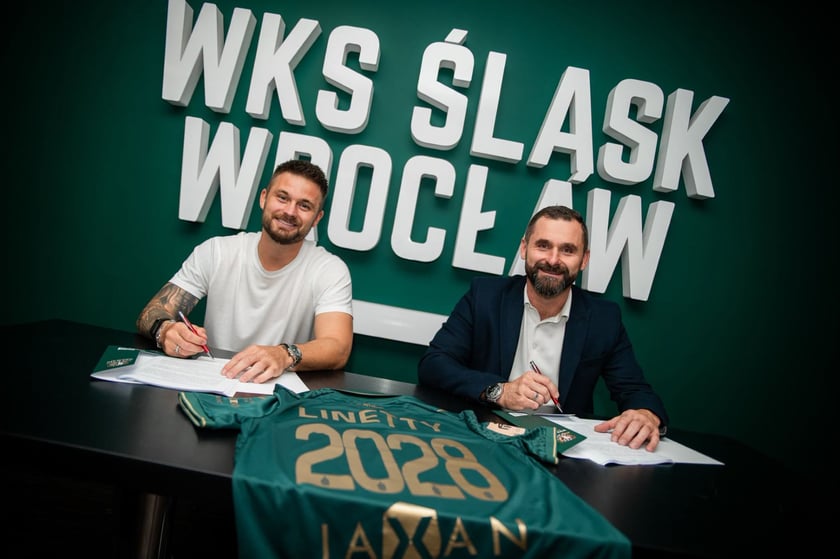 Karol Linetty (z lewej) i Remigiusz Jezierski, prezes Śląska Wrocław (z prawej) siedzą przy stole na którym leży koszulka klubowa, w tle na ścianie biały napis "WKS Śląsk Wrocław"..