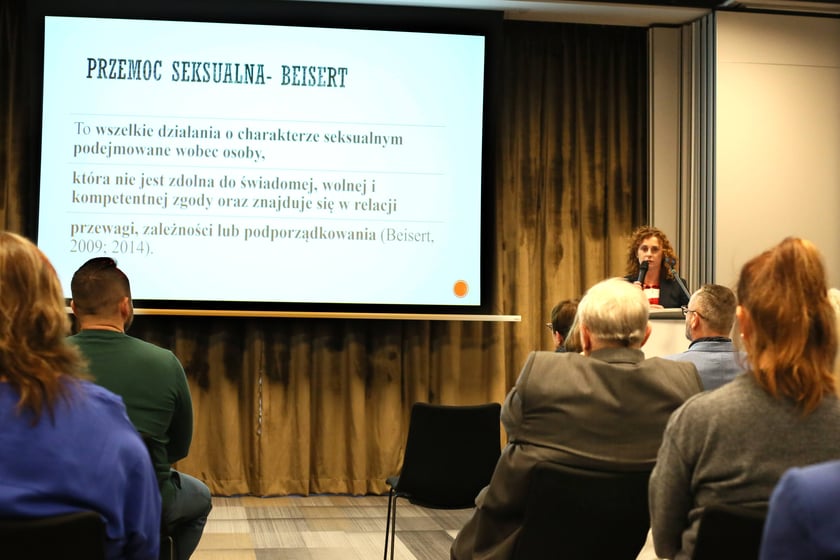 Kobieta prowadząca prezentację na konferencji naukowej, zwrócona do słuchaczy siedzących na sali. Na ekranie za nią wyświetlany jest slajd na temat przemocy seksualnej z tekstem teoretycznym.