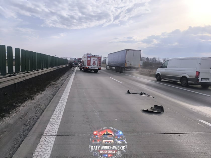 Zdjęcie rozbitych elementów samochodu na jezdni, w tle pojazd straży pożarnej i jadąca ciężarówka.