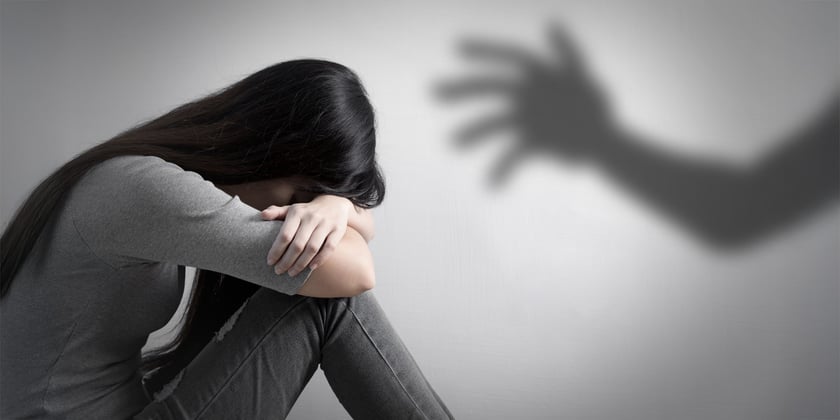 Powiększ obraz: Kobieta siedząca w pozycji przytulonej, z głową pochyloną na kolanach. Na tle widoczny rozmyty cień uniesionej ręki. Czarno-białe zdjęcie sugerujące rozpacz, samotność lub przygnębienie.