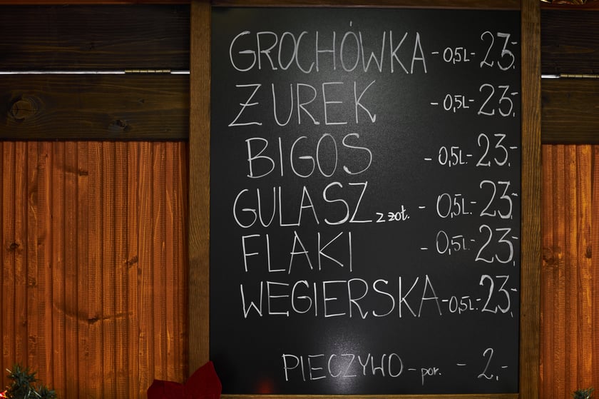 Zdjęcia z Jarmarku Bożonarodzeniowego we Wrocławiu. Na zdjęciu widać tablicę z nazwami potraw i cenami - np. żurek, flaki, grochówka - cena 23 złote
