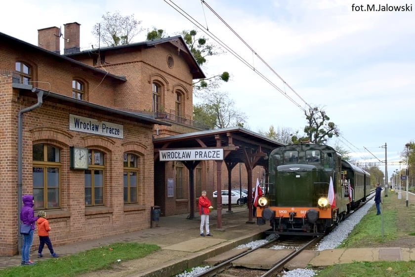 Na zdjęciu widać pociąg retro, który stoi na stacji Wrocław Pracze. Po lewej stronie zdjęcia widać budynki stacji kolejowej z napisem Wrocław Pracze, po prawej - na peronie - zabytkowy  pociąg