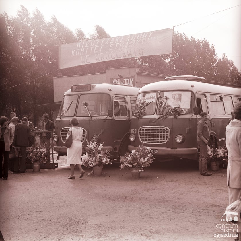 Na zdjęciu dwa autobusy jelcz 043 w bramie wjazdowej do zajezdni autobusowej nr VII. Przed nimi dużo kwiatów, a w górze baner z napisem „Międzyzakładowy Komitet Strajkowy we Wrocławiu”.