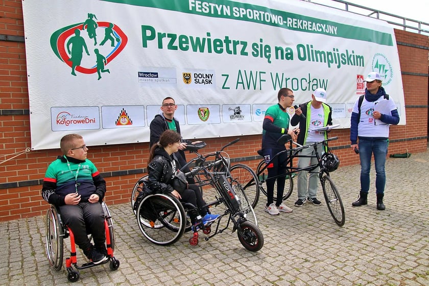 Sześć os&oacute;b przed banerem z napisem "Przewietrz się na Olmpijskim". Kolejno: dwie osoby siedzą na w&oacute;zku inwalidzkim, dwie stoją przed rowerami, dwie stoją obok. Zdjęcia z festynu z 2019 r.