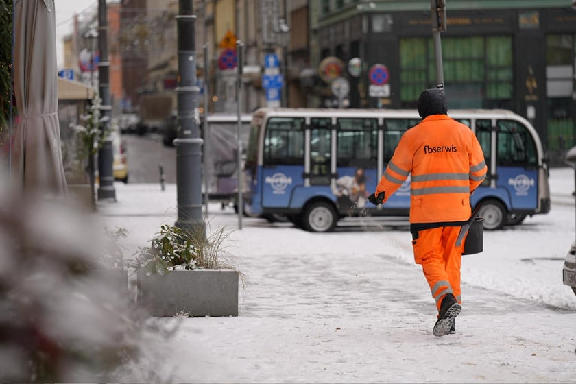 Pierwszy śnieg we Wrocławiu, 24.11.2025. Pracownik z pomarańczowym kombinezonie posypujący solą chodnik.