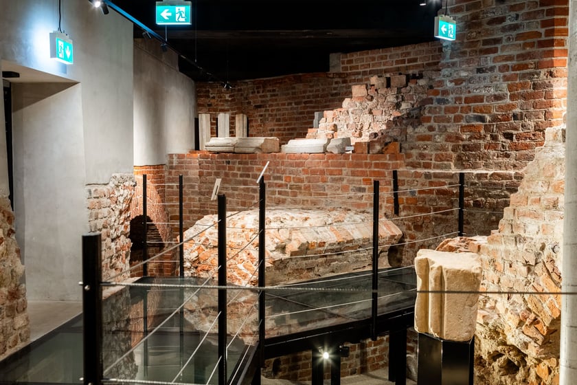 Na pierwszym planie XII wieczny detal kamieniarski pochodzący z obiektu sakralnego; w tle ściana fundamentowa 8 bocznej auli Henryka IV Probusa