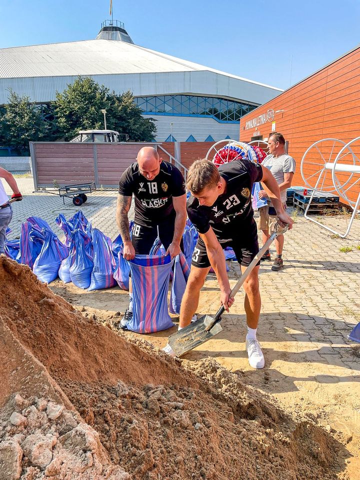 Piłkarze ręczni Śląska (ubrani w klubowe stroje) nasypują łopatami piach do worków i wrzucają je na naczepę