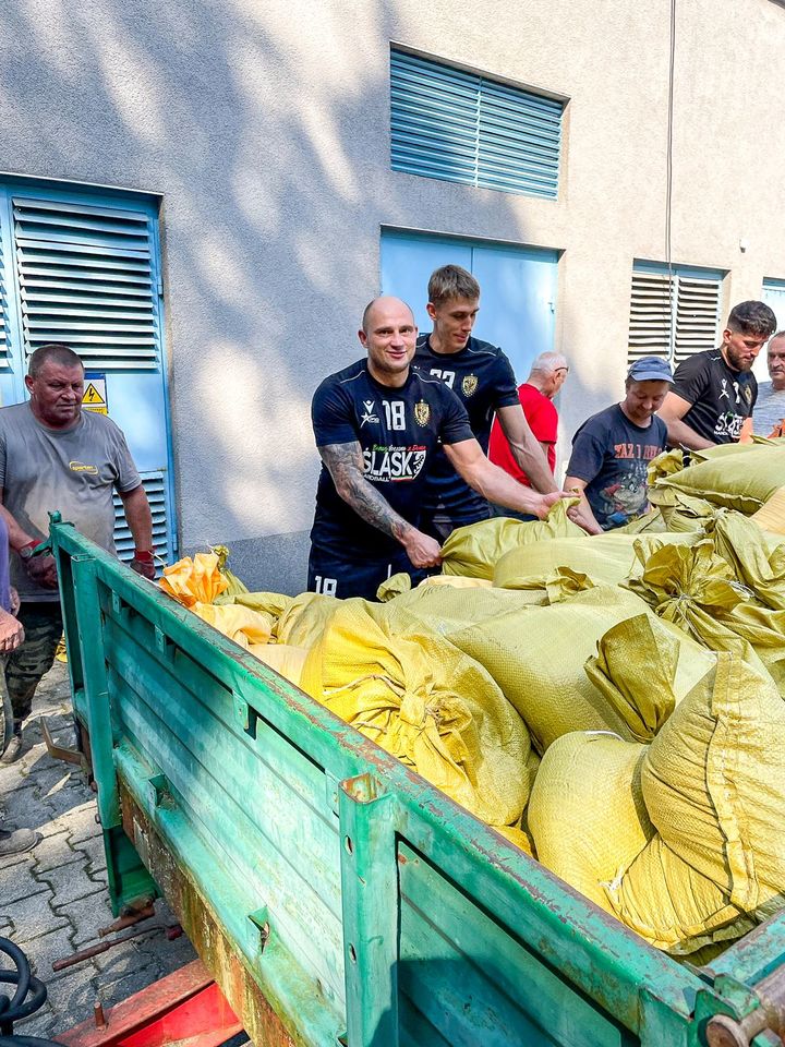 Piłkarze ręczni Śląska (ubrani w klubowe stroje) nasypują łopatami piach do worków i wrzucają je na naczepę