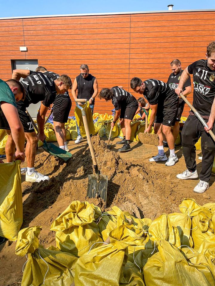 Piłkarze ręczni Śląska (ubrani w klubowe stroje) nasypują łopatami piach do worków i wrzucają je na naczepę
