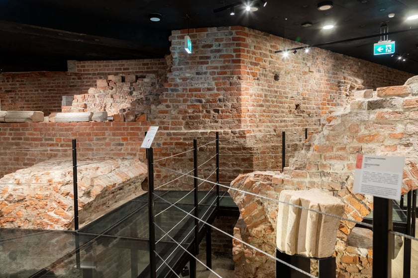 Na pierwszym planie XII wieczny detal kamieniarski pochodzący z obiektu sakralnego; w tle ściana fundamentowa 8 bocznej auli Henryka IV Probusa
