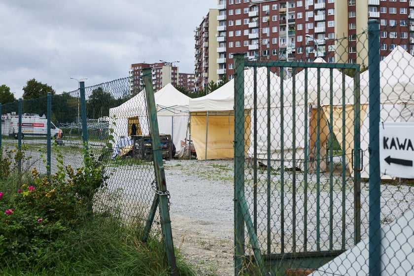 Chodnik osiedlowy. Przy nim ogrodzony teren placu targowego. Otwarta furtka - wejście na teren targowiska. Stanowiska handlowe w postaci białych namiotów. W tle zabudowania osiedla i duże bloki.