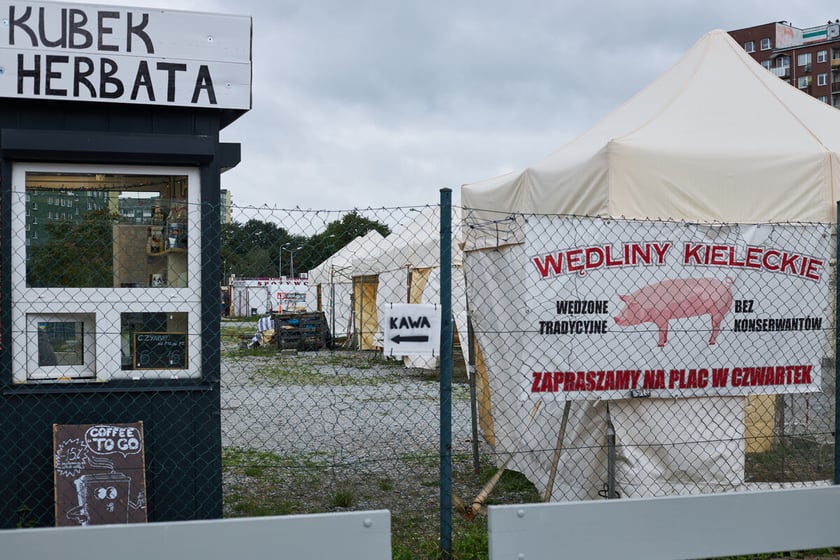 Ogrodzony teren placu targowego. Na ogrodzeniu reklamy sprzedawców. Stanowiska handlowe. Białe namioty.