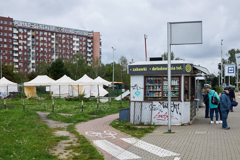 Na pierwszym planie kiosk i przystanek tramwajowy. Na lewo ścieżka rowerowa i teren zieleni. W tle widać ogrodzony teren placu targowego i białe namioty oraz zabudowania osiedla i duże bloki.