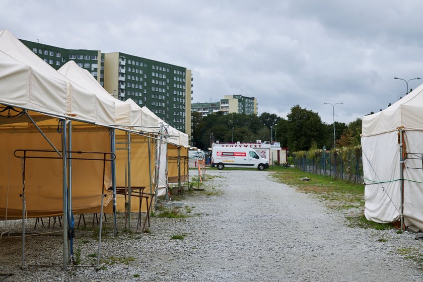 Teren targowiska. Biała, żwirowa nawierzchnia. Po lewej i prawej stronie stanowiska handlowe - białe namioty. W tle biały samochód dostawczy.