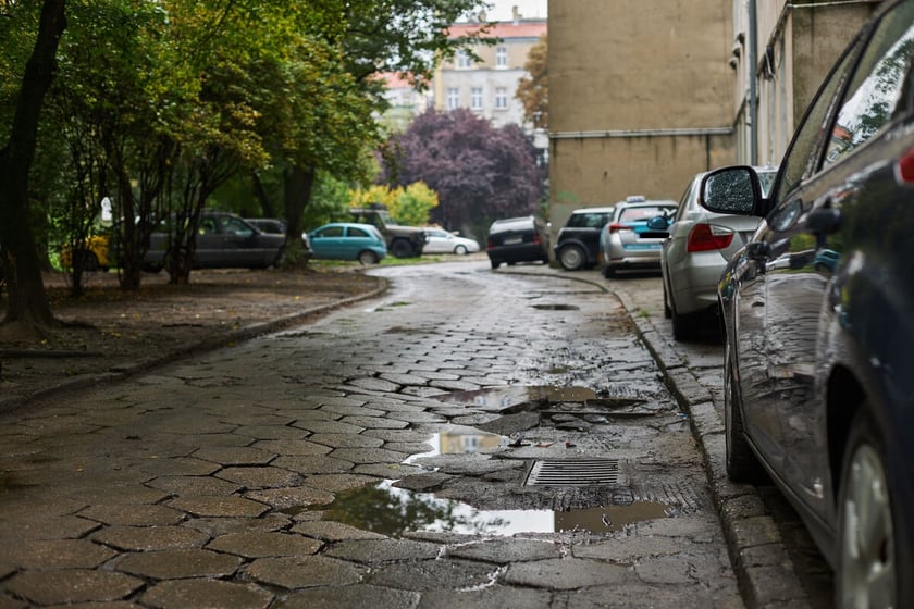 Zaniedbana, dziurawa, podwórkowa ulicę z betonowych płyt. Po prawej przy budynku parkujące w samochody. Po lewej skwer i parkujące wśród drzew samochody.