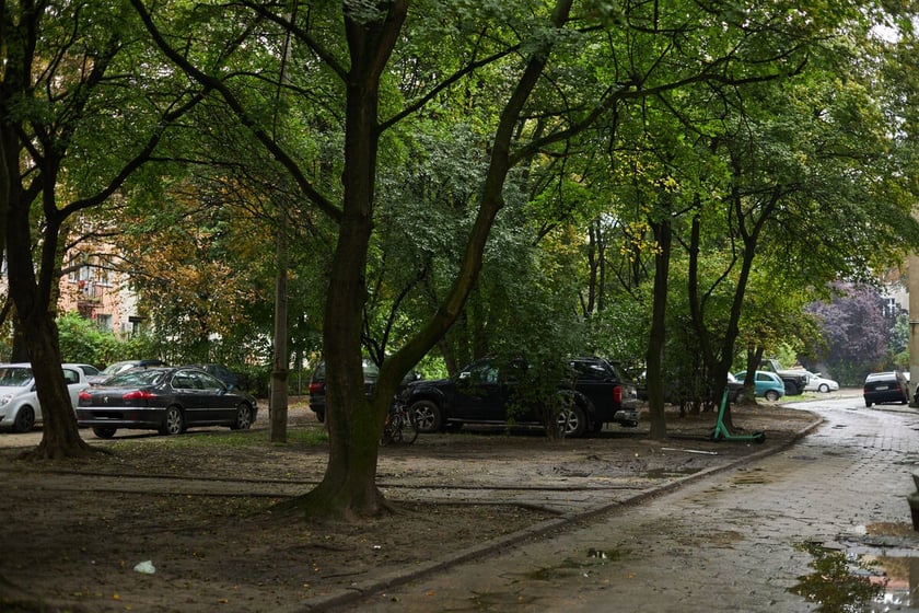 Po prawej stronie ulica podwórkowa.  Skwer i teren zieleni. Parkujące na nim samochody. W tle zabudowania mieszkalne.
