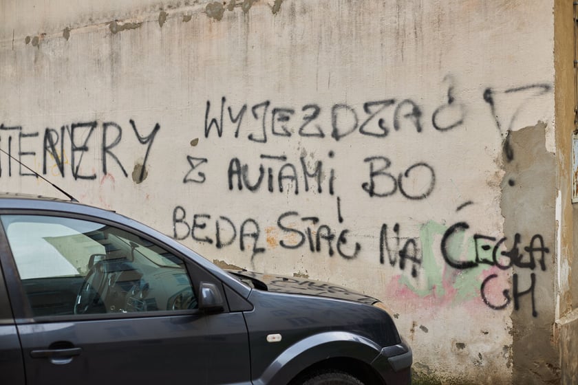 Na zdjęciu bok zaparkowanego pod ścianą samochodu. Na ścianie napis: "wyjeżdżać z autami bo będą stały na cegłach"