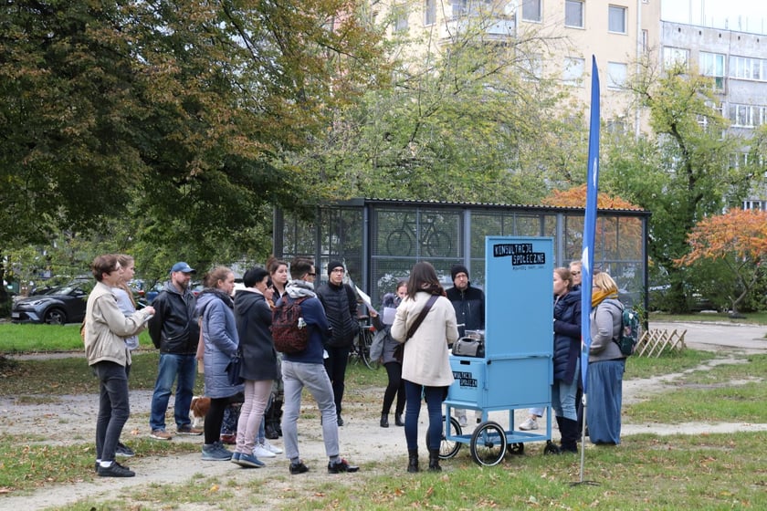 Grupa mieszkańców stoi na podwórku przy wózku konsultacyjnym. W tle wiata rowerowa.