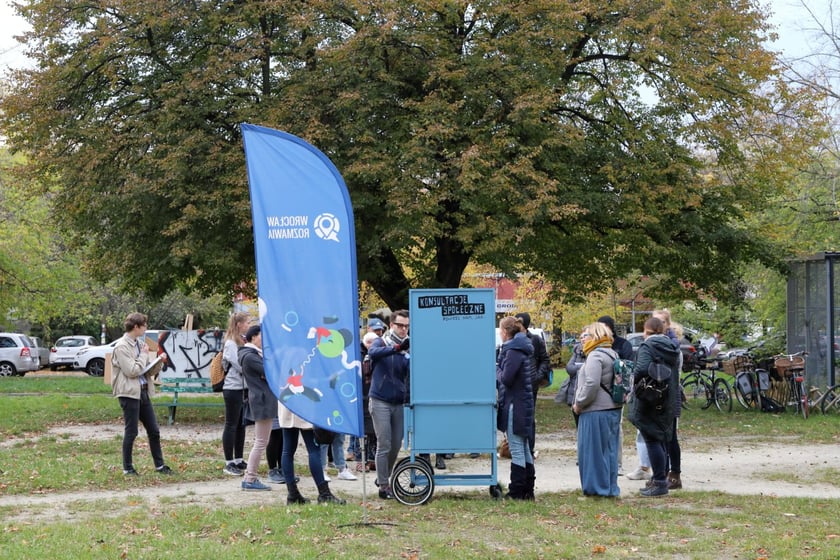 Grupa mieszkańców stoi na podwórku przy wózku konsultacyjnym.