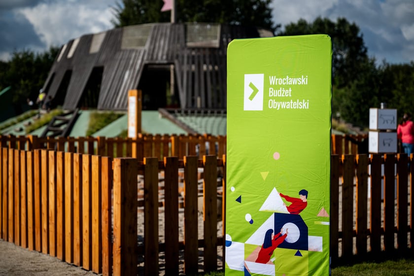 Zielony baner z logotypem Wrocławskiego Budżetu Obywatelskiego (WBO). W tle plac zabaw "Średniowieczny Wrocław" w parku Tysiąclecia - zrealziowany dzięki głosom oddanym w WBO.