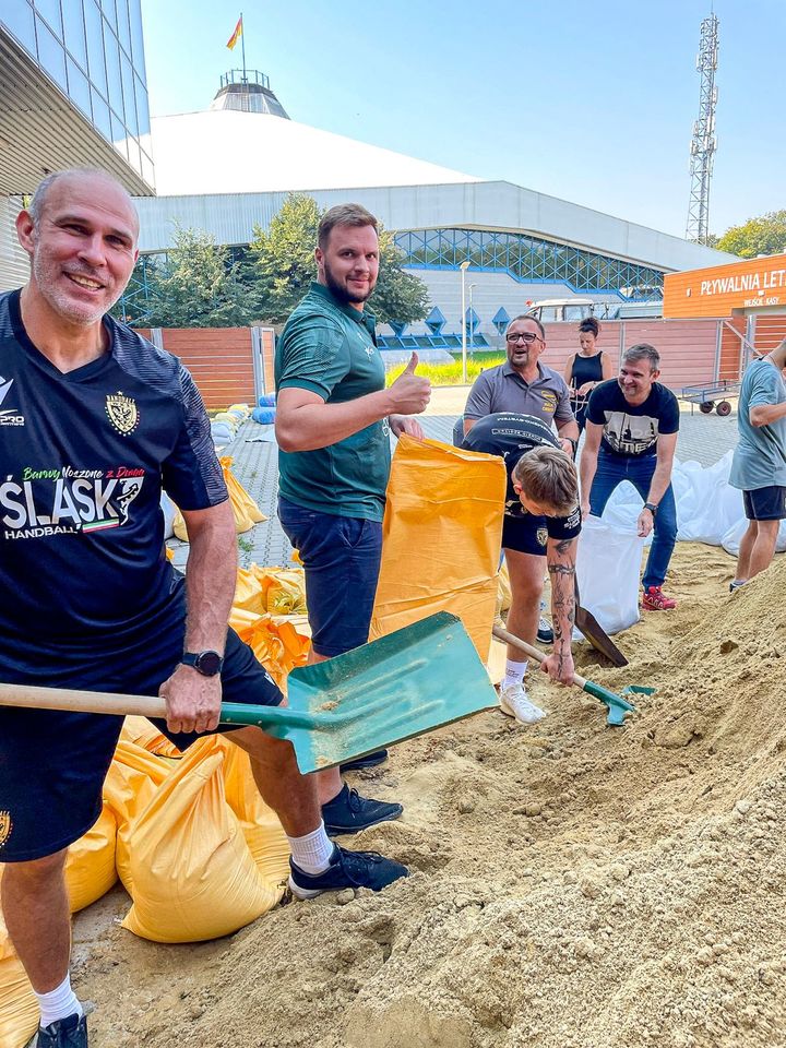 Piłkarze ręczni Śląska (ubrani w klubowe stroje) nasypują łopatami piach do worków i wrzucają je na naczepę