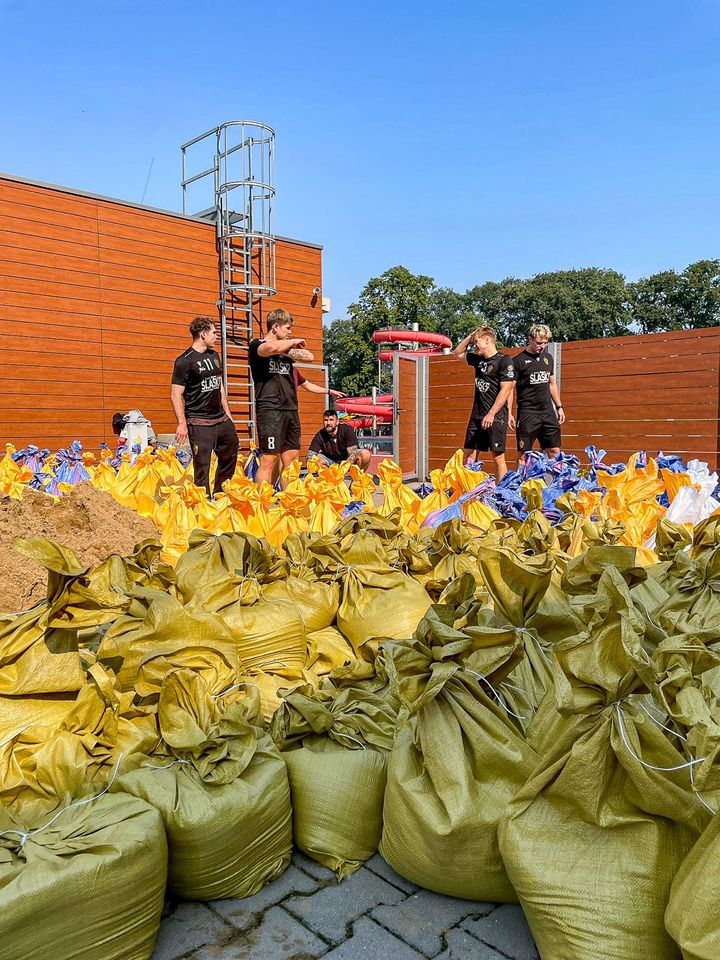 Piłkarze ręczni Śląska (ubrani w klubowe stroje) nasypują łopatami piach do worków i wrzucają je na naczepę