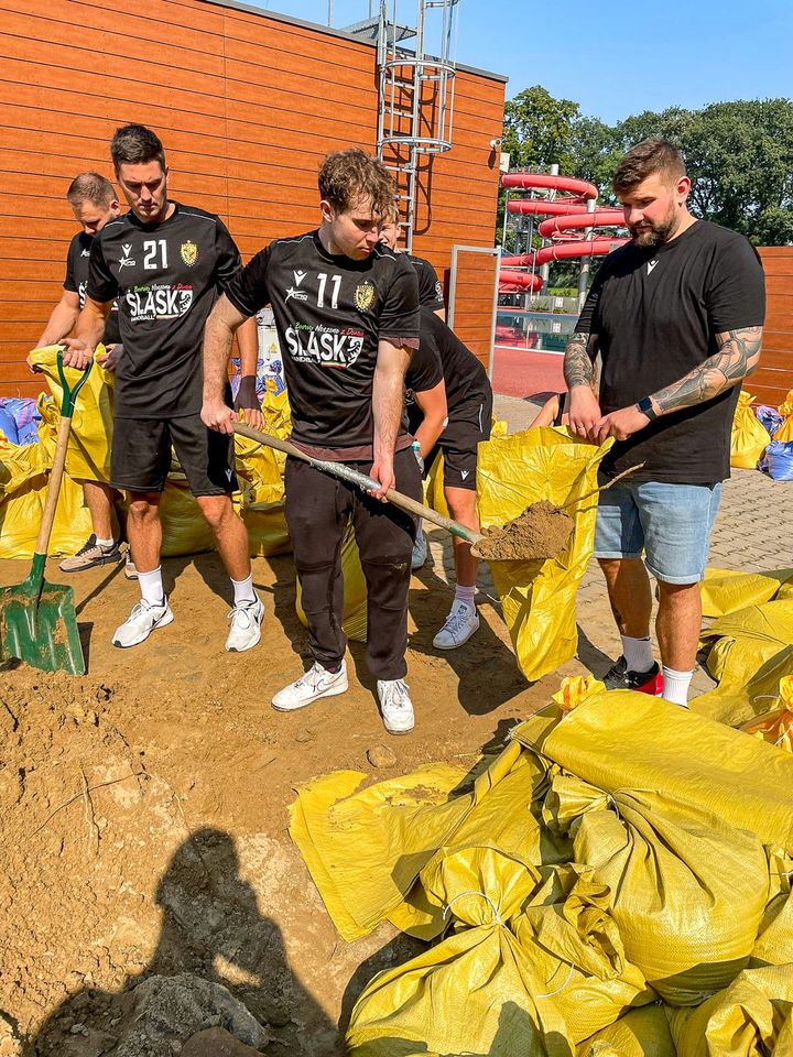 Piłkarze ręczni Śląska (ubrani w klubowe stroje) nasypują łopatami piach do worków i wrzucają je na naczepę