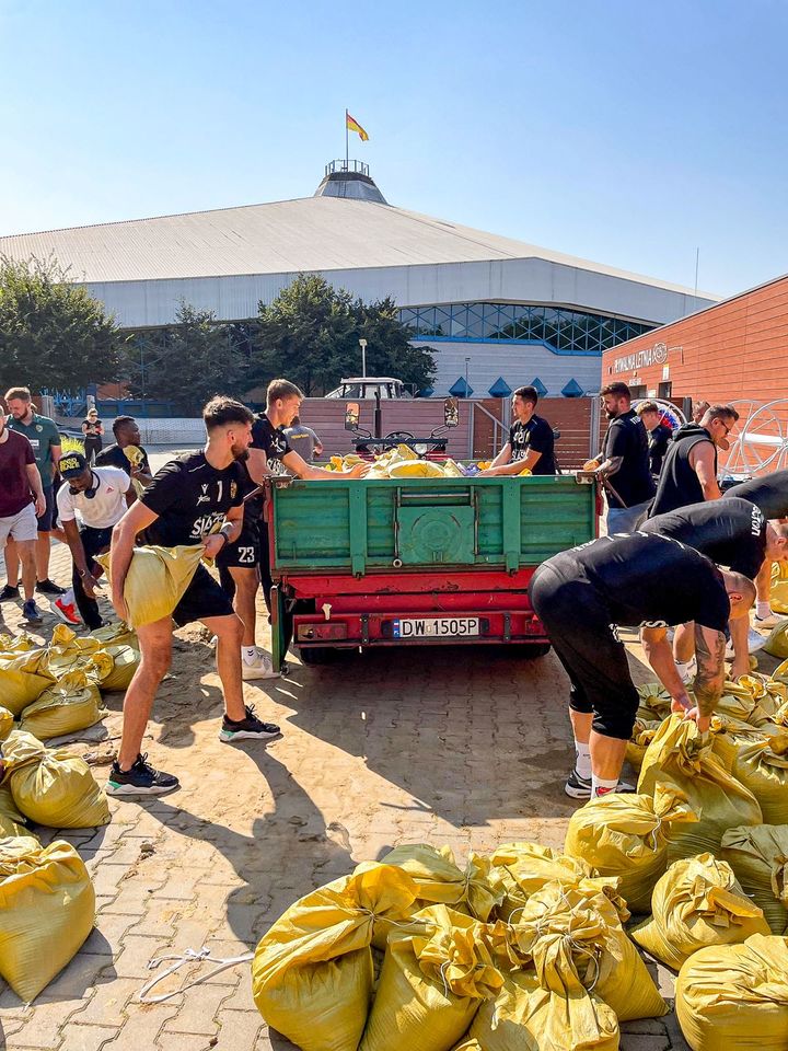Piłkarze ręczni Śląska (ubrani w klubowe stroje) nasypują łopatami piach do worków i wrzucają je na naczepę