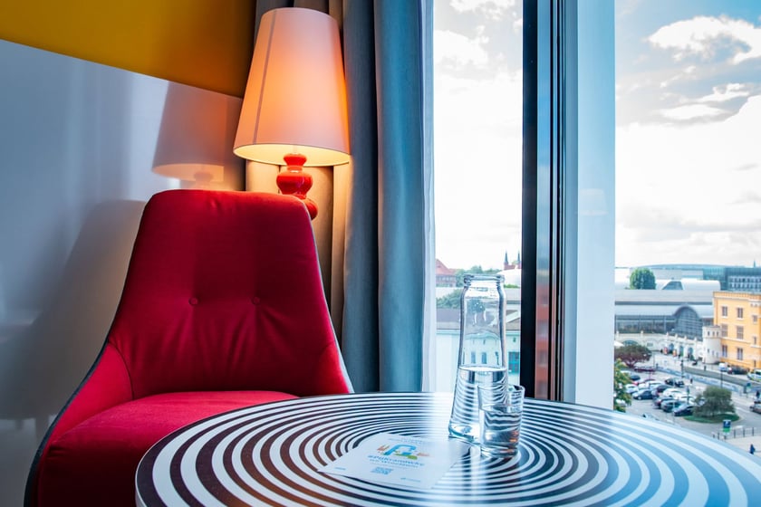 Fotel i stolik przy oknie w pokoju hotelowym, na stoliku stoi szklanka i karafka z wodą.