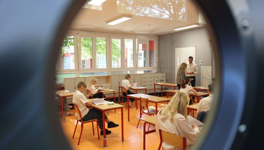 Przez drzwi widać uczniów siedzących w klasie w ławkach 