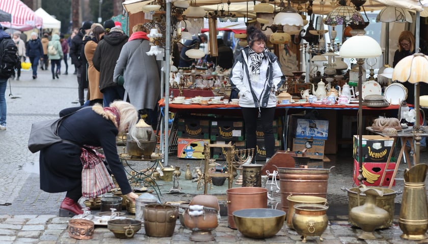 Na zdjęciu, na pierwszym planie, widać rzeczy, które można było kupić na targu staroci przed Halą Stulecia we Wrocławiu. W tle stoją kupujący ludzie i panią, która sprzedaje starocie 