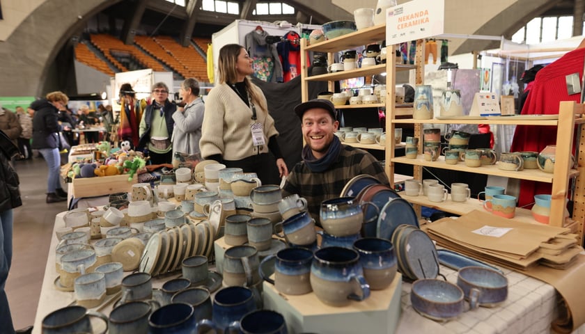 Na zdjęciu, na pierwszym planie, widać ceramikę - kubki i talerze, które są wystawione na sprzedaż na targach w Hali Stulecia. W centralnym miejscu zdjęcia siedzi uśmiechnięty mężczyzna - sprzedawca ceramiki 
