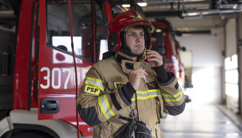 Na zdjęciu st. str. Mateusz Zieliński z Jednostki Ratowniczo-Gaśniczej PSP przy ul. Kosmonautów we Wrocławiu. Mężczyzna jest w strażackim uniformie, w tle widać czerwone wozy strażackie 