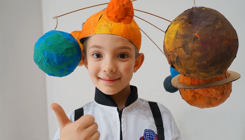 Na zdjęciu chłopiec w pomarańczowym nakryciu głowy z kolorowymi planetami z papieru