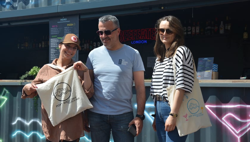 Dziewczyna w czapce z daszkiem trzyma torbę z logo i napisem Port Węglowy Beach Bar, obok mężczyzna w okularach słonecznych i kobieta w pasiastej bluzce