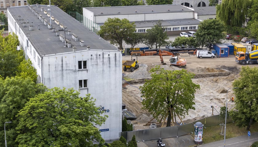 Komisariat Wrocław-Stare Miasto, korony drzew, pojazdy i ludzie na rozkopanej, ogrodzonej działce