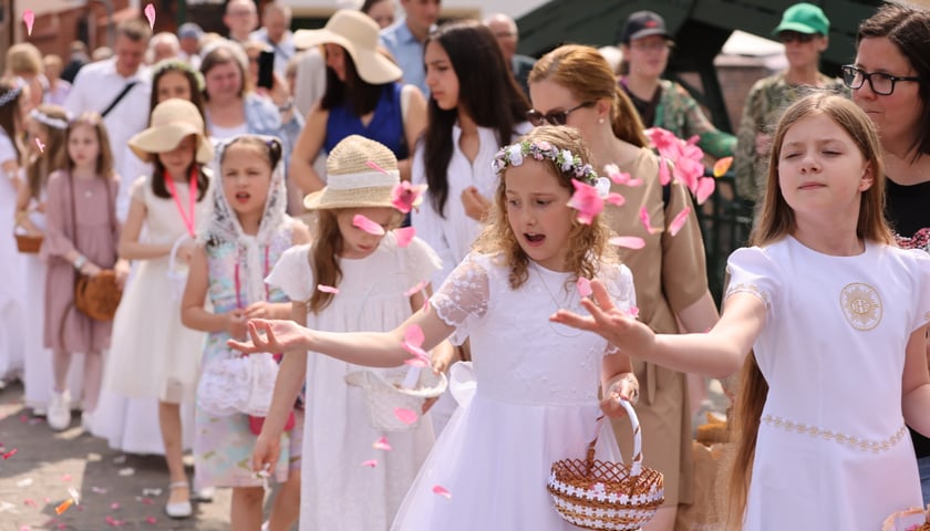 Procesja Bożego Ciała 2023 we Wrocławiu. Dziewczynki w białych komunijnych sukienkach sypią płatki kwiatów, które niosą w koszyczkach. Za nimi stoją dorośli.