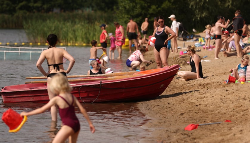 <p>Kąpielisko Glinianki. Grupa os&oacute;b na plaży w strojach kąpielowych, widać też czerwoną ł&oacute;dź, dziecko z czerwonym wiaderkiem weszło do wody</p>