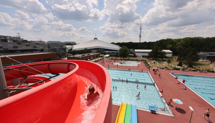 Czerwona zjeżdżalnia na kąpielisku Orbita. Widok z góry: zjeżdżające dziecko, na dole baseny i kąpiący się ludzie oraz druga zjeżdżalnia - żółto-zielono-niebieska