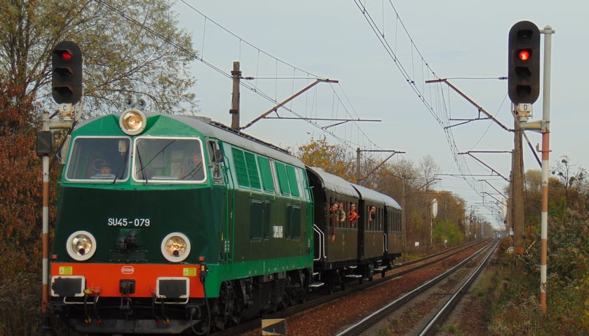 Historyczny pociąg z wagonami jedzie na torach, z okien wychylają się pasażerowie