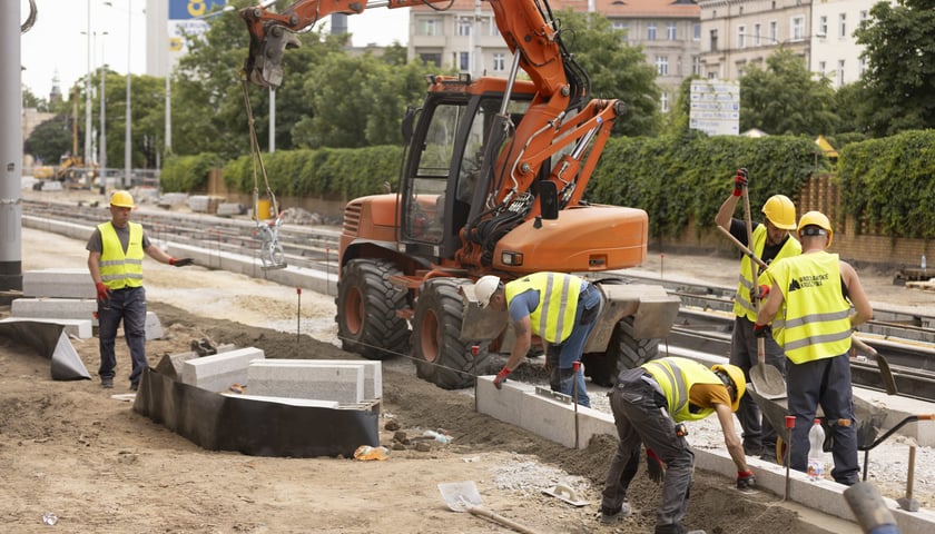 Piątka robotników, w żółtych kamizelkach, układa chodnik na placu przebudowy ulicy Pomorskiej i Staszica we Wrocławiu.