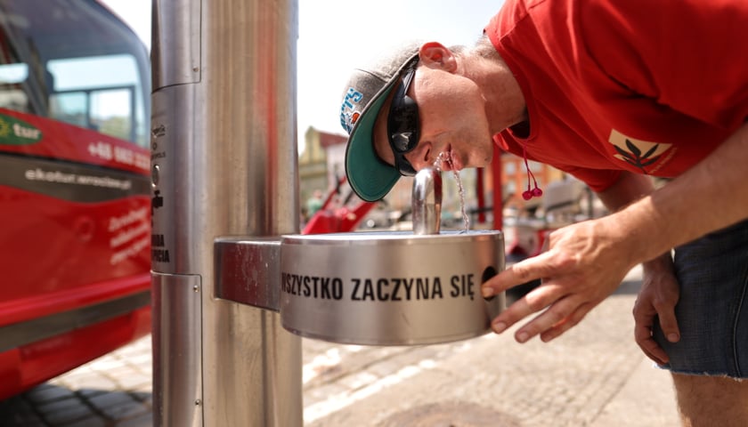 Na zdjęciu mężczyzna w czerwonej koszulce pije wodę ze zdroju ulicznego