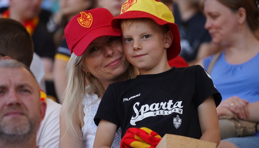 Na trybunach stadionu matka w czerwonej czapce z daszkiem i siedzącym na jej kolanach synkiem w koszulce Betardu i żółto-czerwonym kapelusiku 