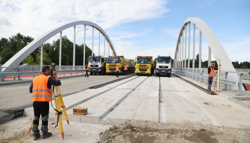 Technik w pomarańczowej kamizelce patrzy przez przyrząd pomiarowy na ciężarówki stojące na moście Chrobrego