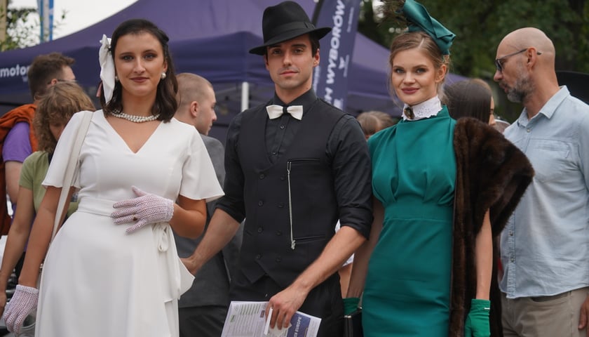 Tysiące widzów na Wielkiej Wrocławskiej na torze wyścigów konnych Partynice. Na zdjęciu dwie młode kobiety i młody mężczyzna w eleganckich strojach.
