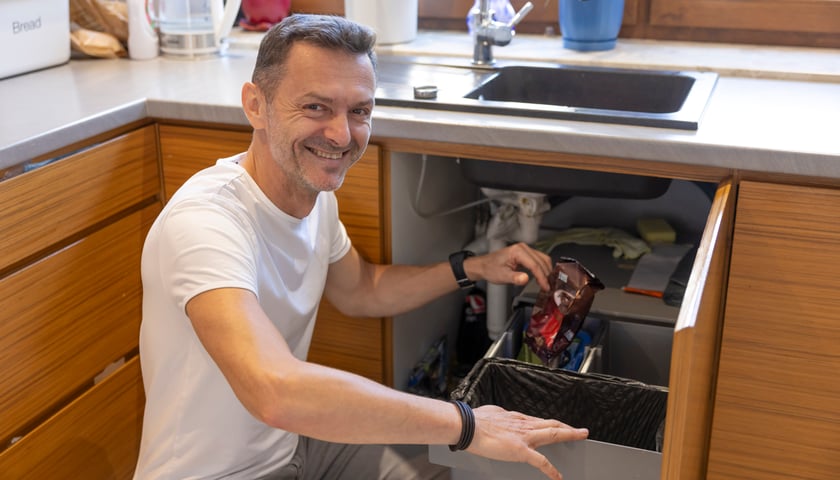 Kuchnia, mężczyzna - Krystian Sroka - w białej koszulce kuca przy otwartej szafce pod zlewem i pokazuje znajdujące się tam pojemniki na odpady 