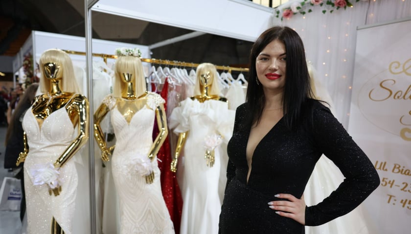 Targi Ślubne w Hali Stulecia. Młoda kobieta w czarnej sukience, z tyłu stoją białe suknie ślubne