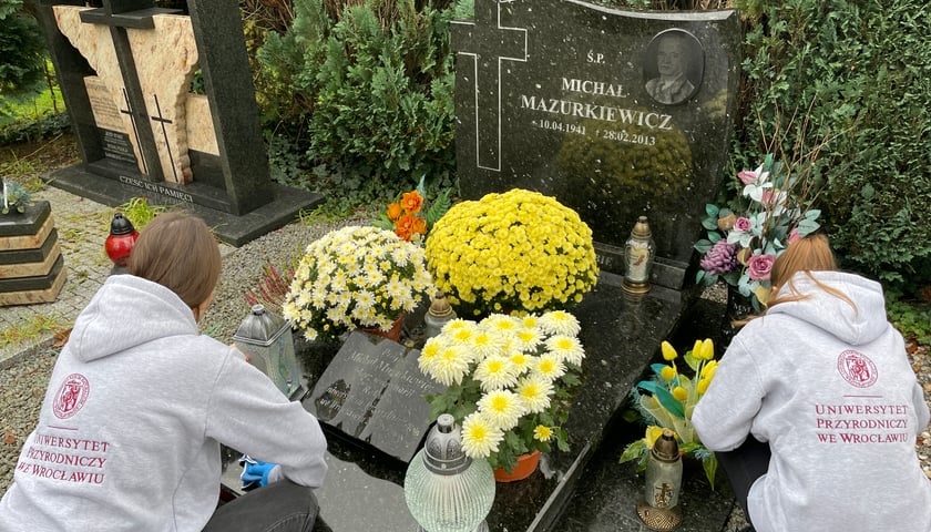 Dwie osoby odwrócone plecami kucają obok grobu. Na płycie leżą żółte kwiaty 
