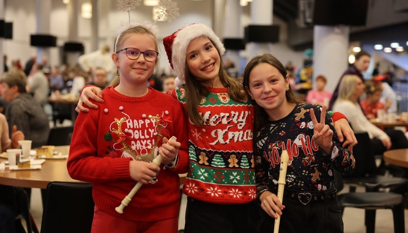 Trzy dziewczynki ubrane w swetry ze świątecznymi motywami,  dwie trzymają w dłoni flety